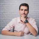 Positive young male in shirt with wristwatch sitting at desk with hand at chin while smiling and looking at camera in room with white brick wall